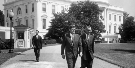 President John F. Kennedy and Vice President Lyndon B. Johnson walk across the South Lawn driveway at the White House in Washington, D.C. to attend a ceremony marking the 50th anniversary of the first state Workmen's Compensation Law, enacted in Wisconsin in 1911. Secret Service Agent Bob Lilley follows.